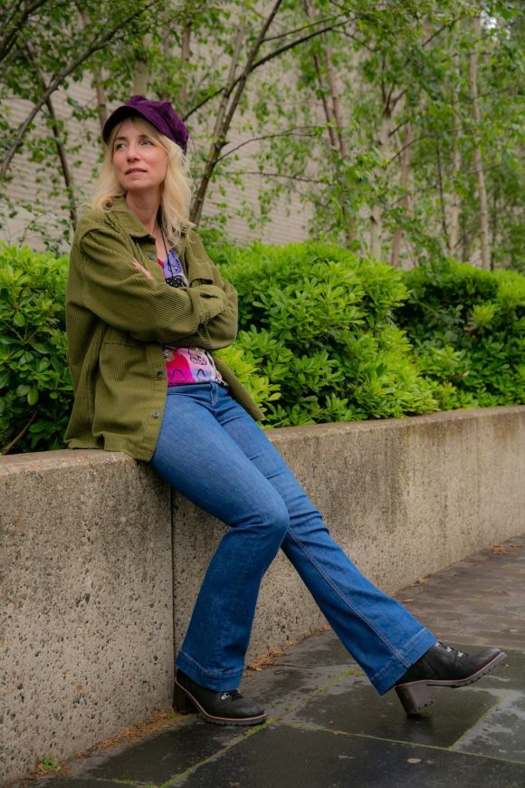 Freelance instructional design expert sits on concrete wall in jeans, a green jacket and purple hat